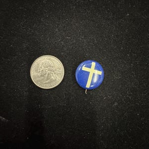 May include: A silver quarter coin next to a blue button with a yellow cross design. The button has a pin on the back. Both items are on a black surface.