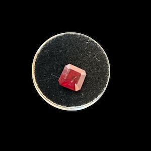 May include: A faceted, rectangular-cut, deep red gemstone displayed on a small, round, black velvet surface. The gemstone is the focal point, set against a stark black background, highlighting its color and clarity.
