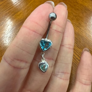 May include: A silver belly button ring featuring a top ball and a dangling heart-shaped charm. The charm has a light blue gemstone set within a heart-shaped frame. The design includes a smaller heart-shaped charm below.