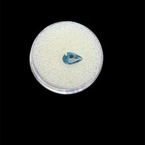 May include: A pear-shaped, light blue gemstone displayed in a clear plastic container with a white foam insert. The gemstone has a polished surface and is set against a black background.