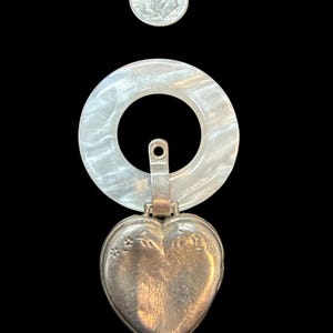 May include: A vintage silver heart-shaped locket pendant with floral detailing, suspended from a pearlescent, circular charm. A US dime is included for size reference. The locket is likely made of sterling silver.