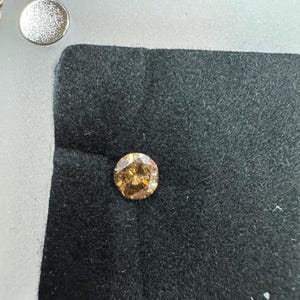 May include: A round, light brown diamond set on a black velvet surface, within a silver-colored frame. The diamond's facets reflect light, showcasing its brilliance. The setting includes a small, silver-colored circular element.