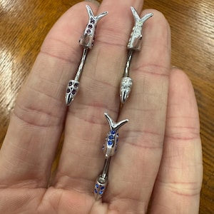 May include: Three silver industrial barbells with decorative ends. Each barbell features a design with a pointed tip and a stylized top, adorned with either purple or blue gemstones. The jewelry is held in a hand.