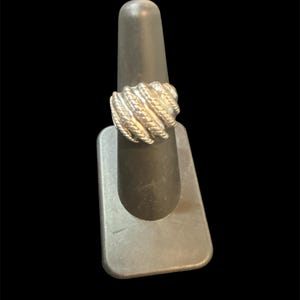 May include: Silver-toned ring with a textured, rope-like design. The ring is displayed on a black ring holder against a black background. The ring's design features multiple bands of twisted metal, creating a bold and eye-catching look.