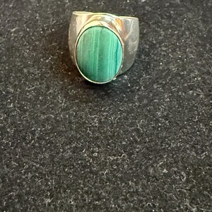 Puede incluir: Un anillo de plata con una gran piedra ovalada de malaquita verde. La piedra está engastada en un bisel y tiene un patrón de rayas. El anillo tiene una banda ancha.