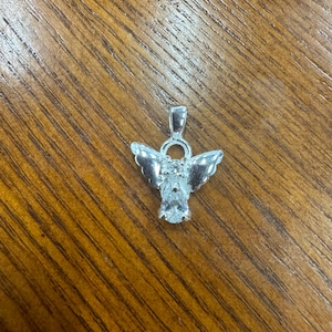 May include: A silver-toned angel pendant with a clear gemstone body and detailed wings. The pendant has a loop at the top for a chain. The angel is set against a wooden background.