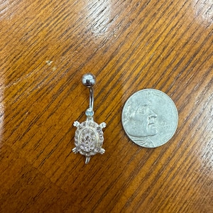 May include: A silver belly button ring with a turtle design. The turtle has pink and clear gemstones. The ring is next to a quarter coin on a wooden surface.