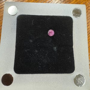 May include: A small, round, pink gemstone set in a black velvet display within a clear, rectangular frame. The frame has four silver-colored circular magnets at each corner. The gemstone is the focal point.