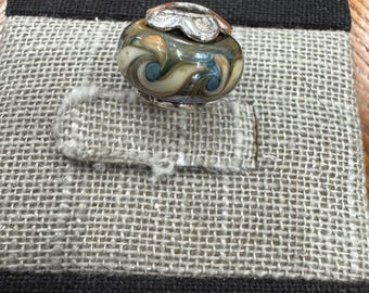 Puede incluir: Una cuenta decorativa de vidrio con una base y una parte superior de metal plateado. La cuenta presenta remolinos de colores azul, marrón y crema. La cuenta se muestra sobre una superficie gris similar al lino con un borde negro.