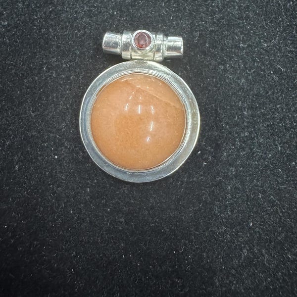 May include: A silver pendant with a circular, peach-colored stone set in a silver bezel. A small, rectangular silver bar with a red gemstone is attached at the top. The pendant is displayed on a black surface.