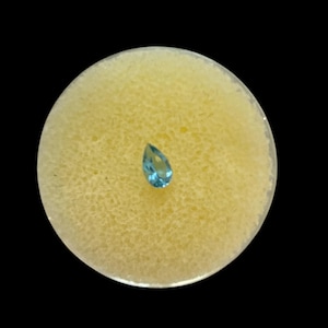 May include: A teardrop-shaped, light blue gemstone is centered on a round, light yellow surface. The gemstone is set against a contrasting black background. The image is a close-up, highlighting the gem's clarity and color.