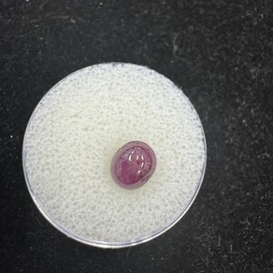 May include: A polished, oval-shaped, deep red gemstone, likely a ruby, displayed in a small, round, clear plastic container with a white, textured interior. The gemstone is the focal point, set against a dark background.
