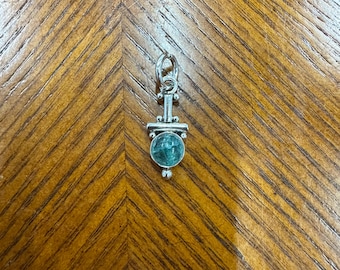 Puede incluir: Un colgante de plata con una piedra redonda de color turquesa. El colgante tiene un diseño decorativo con un lazo en la parte superior para una cadena. El colgante se muestra sobre una superficie de madera.