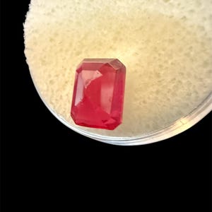 May include: A faceted, rectangular-cut, vibrant red gemstone. The gem is set against a white, textured background, and enclosed in a clear, circular container. The gemstone's colour is a deep, saturated red, with visible internal clarity.