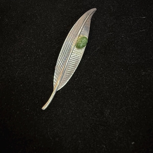 May include: A silver-toned feather-shaped brooch with detailed veining. A small, round, green gemstone is set near the center of the feather. The brooch is displayed against a dark background, highlighting its metallic sheen.