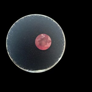 May include: A faceted, round, pink gemstone is centered on a dark gray, circular base. The base has a clear, slightly textured edge. The background is solid black, providing contrast to the gemstone and base.