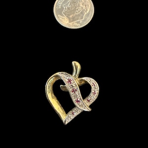 May include: A gold-toned heart-shaped pendant with a ribbon design, adorned with small red and clear gemstones. A U.S. dime is included for size reference. The pendant is set against a black background.