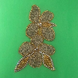 Pode incluir: Uma aplicação floral dourada e com contas com três grupos de flores. As pétalas são cobertas por pequenas contas brilhantes, com contas douradas delineando as bordas. Strass transparentes são colocados no centro das flores e ao longo das folhas. O fundo é verde liso.