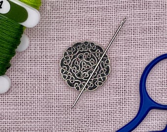 May include: A round silver-toned metal needle minder featuring a black intricate filigree pattern, accompanied by a long sewing needle resting across its surface, positioned near green embroidery floss and blue scissors.