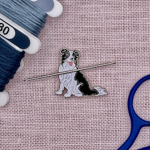 May include: A black, white, and silver enamel pin of a Border Collie dog with a needle passing through it. The dog is sitting with its tongue out. Blue thread and scissors are in the background. The pin is likely for sewing or crafting.