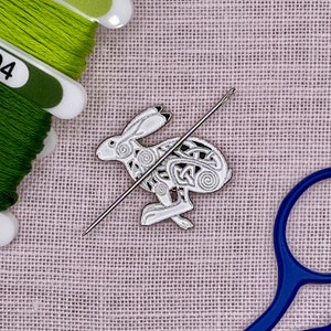 May include: A white enamel hare pin with Celtic knotwork design, a silver needle, and green thread spools. The hare pin is in a running pose. The background is a light purple fabric.