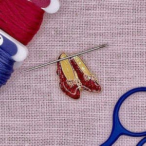 May include: A pair of red glitter enamel shoes with gold detailing, a needle, and spools of red and blue thread. The shoes have a bow detail. The scene is set on a light purple woven fabric, with blue scissors in the corner.