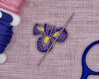 May include: A metal enamel pin shaped like a purple iris flower with yellow accents, featuring a silver-toned needle resting diagonally across the surface of the pin, positioned against a textured light pink fabric background.
