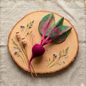 May include: A handcrafted felt beet with green and pink leaves, displayed on a wooden slice. The beet is a deep magenta color, and the leaves have a vibrant green and pink pattern. Small sprigs of dried flowers and grasses are scattered around the beet.