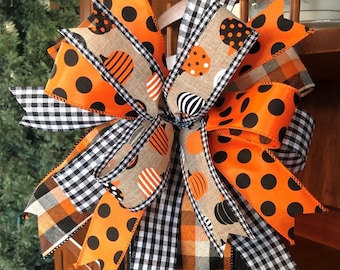 May include: A black lantern with a large orange, black, and white bow. The bow is made of several different ribbons with polka dots, plaid, and pumpkin patterns.