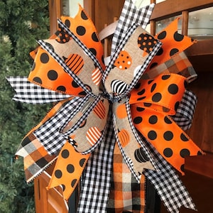 May include: A black lantern with a large orange, black, and white bow. The bow is made of several different ribbons with polka dots, plaid, and pumpkin patterns.