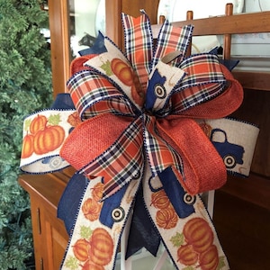 May include: A large, orange, blue, and white plaid bow with a burlap ribbon and a cream ribbon with a pumpkin and truck design. The bow is tied around a white lantern.