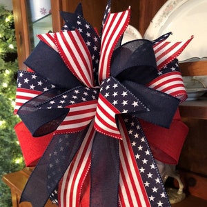 May include: A red, white, and blue patriotic bow made with ribbon featuring the American flag. The bow is tied around a wooden lantern.