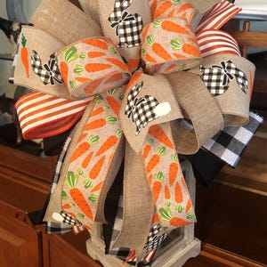 May include: A brown burlap, black and white buffalo check, and orange and white striped ribbon bow with a carrot and bunny print ribbon. The bow is tied around a white lantern.