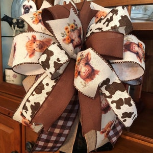 May include: A brown, white, and black plaid bow with a cow print ribbon and a ribbon with a pattern of brown cows with flowers on a white background.