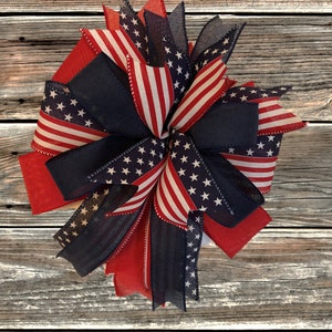 May include: A red, white, and blue patriotic bow with a star-spangled design. The bow is made of ribbon and is tied in a knot. It is a festive decoration for the Fourth of July or other patriotic holidays.