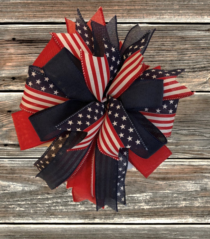 May include: A red, white, and blue patriotic bow with a star-spangled design. The bow is made of ribbon and is tied in a knot. It is a festive decoration for the Fourth of July or other patriotic holidays.
