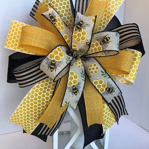 May include: A yellow and black bee-themed bow with a honeycomb pattern and bee illustrations. The bow is tied around a white lantern.