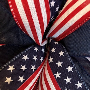 May include: Close-up of a red, white, and blue American flag ribbon. The ribbon is folded in a starburst pattern, with the stars of the flag visible in the center.