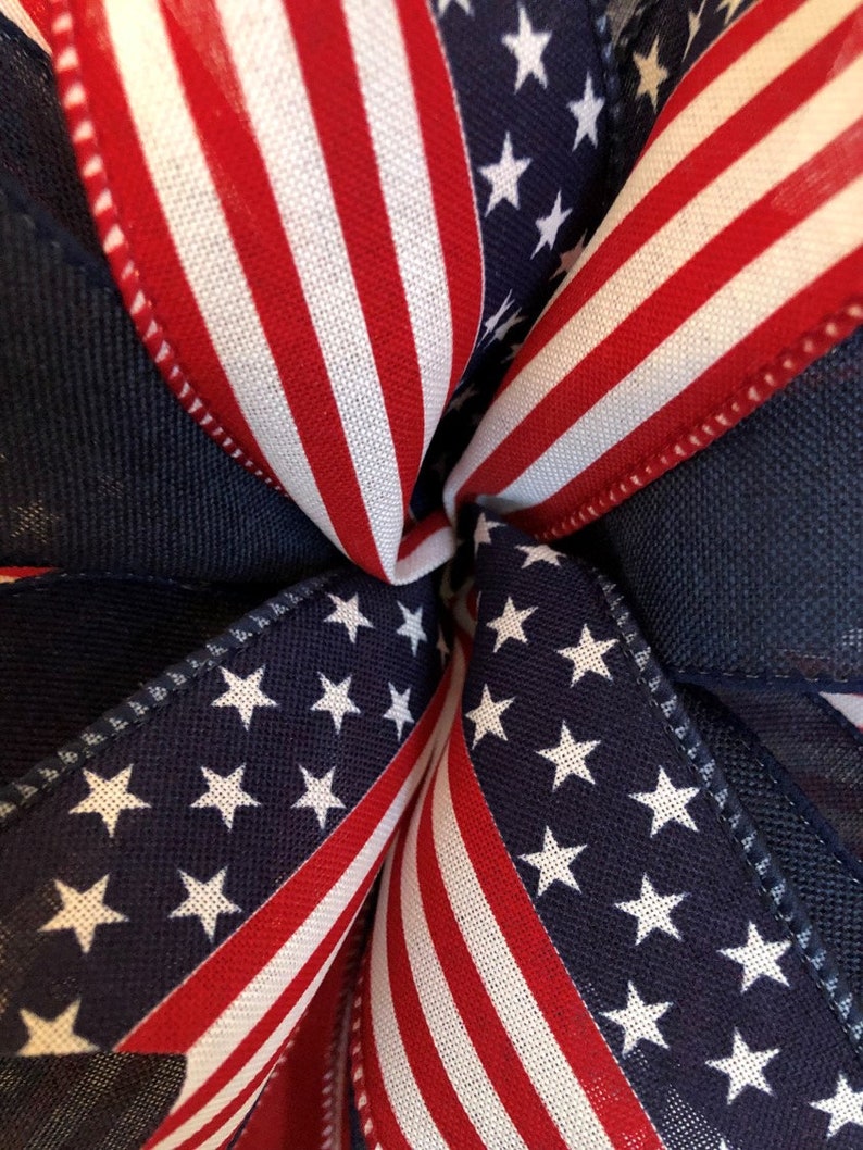 May include: Close-up of a red, white, and blue American flag ribbon. The ribbon is folded in a starburst pattern, with the stars of the flag visible in the center.
