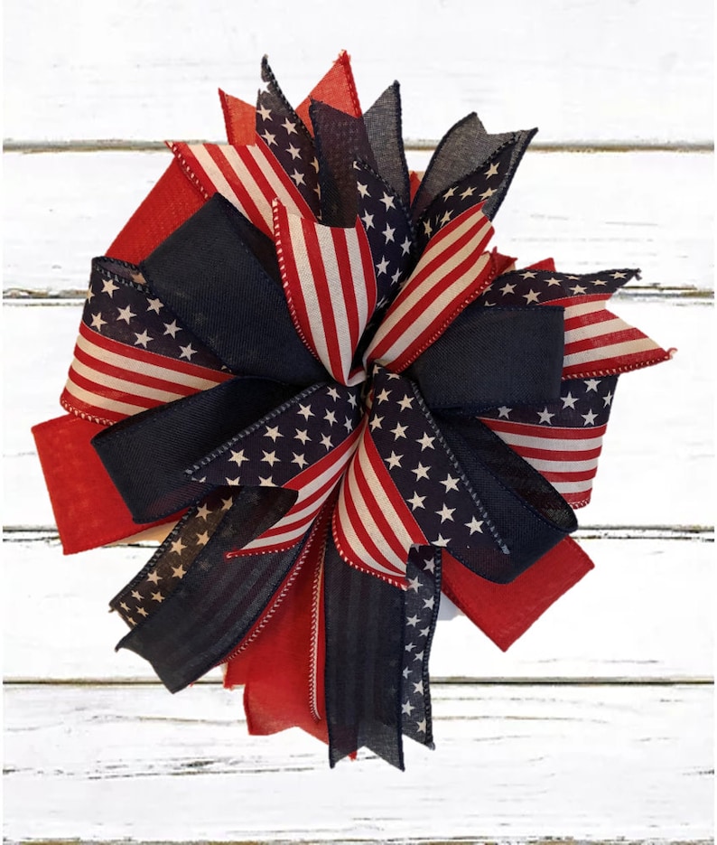 May include: A red, white, and blue patriotic bow with a star-spangled design. The bow is made of ribbon and is perfect for decorating for the Fourth of July or other patriotic holidays.