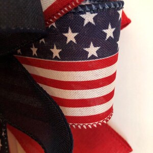 May include: A close-up of a red, white, and blue fabric bow with a star pattern. The bow is made of a woven fabric with a stitched edge.