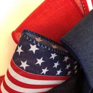 May include: A close-up of a red, white, and blue fabric ribbon with stars and stripes. The ribbon is folded and tied into a bow.
