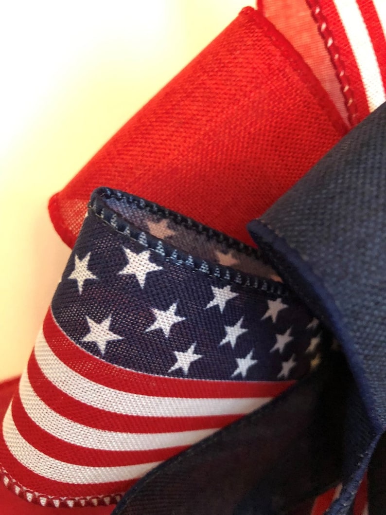 May include: A close-up of a red, white, and blue fabric ribbon with stars and stripes. The ribbon is folded and tied into a bow.
