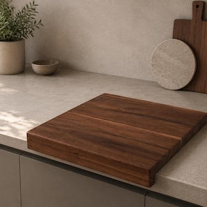 Puede incluir: Una tabla de cortar cuadrada de madera de color marrón oscuro con una superficie lisa y plana y un borde grueso. La tabla está colocada sobre una encimera de color claro, mostrando un patrón de veta de madera visible en toda su estructura.