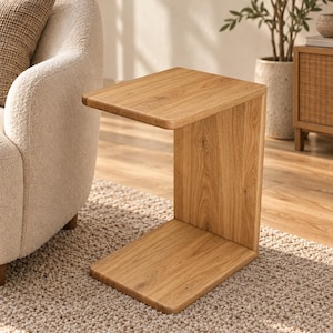 Peut inclure: Une table d'appoint en forme de C fabriquée en bois clair avec un motif de grain visible. La table présente un plateau rectangulaire plat, un panneau de support vertical et une base rectangulaire plate, formant une structure continue.