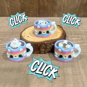 May include: Three colorful miniature mugs with rainbow-colored bodies and white handles. Each mug features a different topping, including whipped cream, marshmallows, and a swirl. The word "CLICK" is displayed in speech bubbles around the mugs.