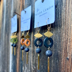 May include: Three pairs of earrings with gold hoops, wood accents, and black lava rock beads. The earrings are hanging from a white card with the text "the WILDE LEMON @thewildelemon".