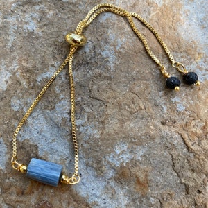 May include: A gold chain bracelet with a blue stone and black lava beads. The bracelet has a sliding clasp.