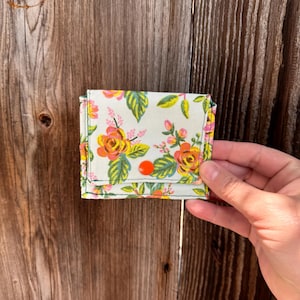 May include: A small, floral-patterned wallet with a light background, featuring yellow and orange roses and green leaves. The wallet is secured with a bright orange button. The edges are trimmed with yellow stitching.