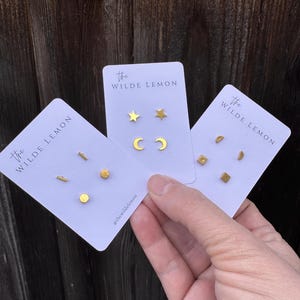 May include: Three cards with gold earrings on them. The cards are white with the text "The Wilde Lemon" on them. The earrings are in the shape of a star, crescent moon, a small rectangle, a small circle, and a small line.